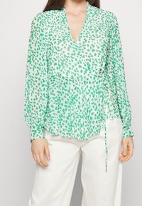 Grüne Bluse mit Muster, V-Ausschnitt, langen Ärmeln, plissee Design und einem Bindeband in der Taille, kombiniert mit weißen Hosen.