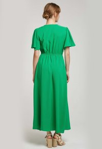 Robe verte, longue jusqu'au sol, avec des manches bouffantes, une taille élastique et une texture douce. Le dos présente un col rond simple et une silhouette fluide.