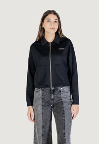 Giacca nera cropped con zip frontale, due tasche anteriori e colletto. Include il logo "GUESS" ricamato in oro sul petto.