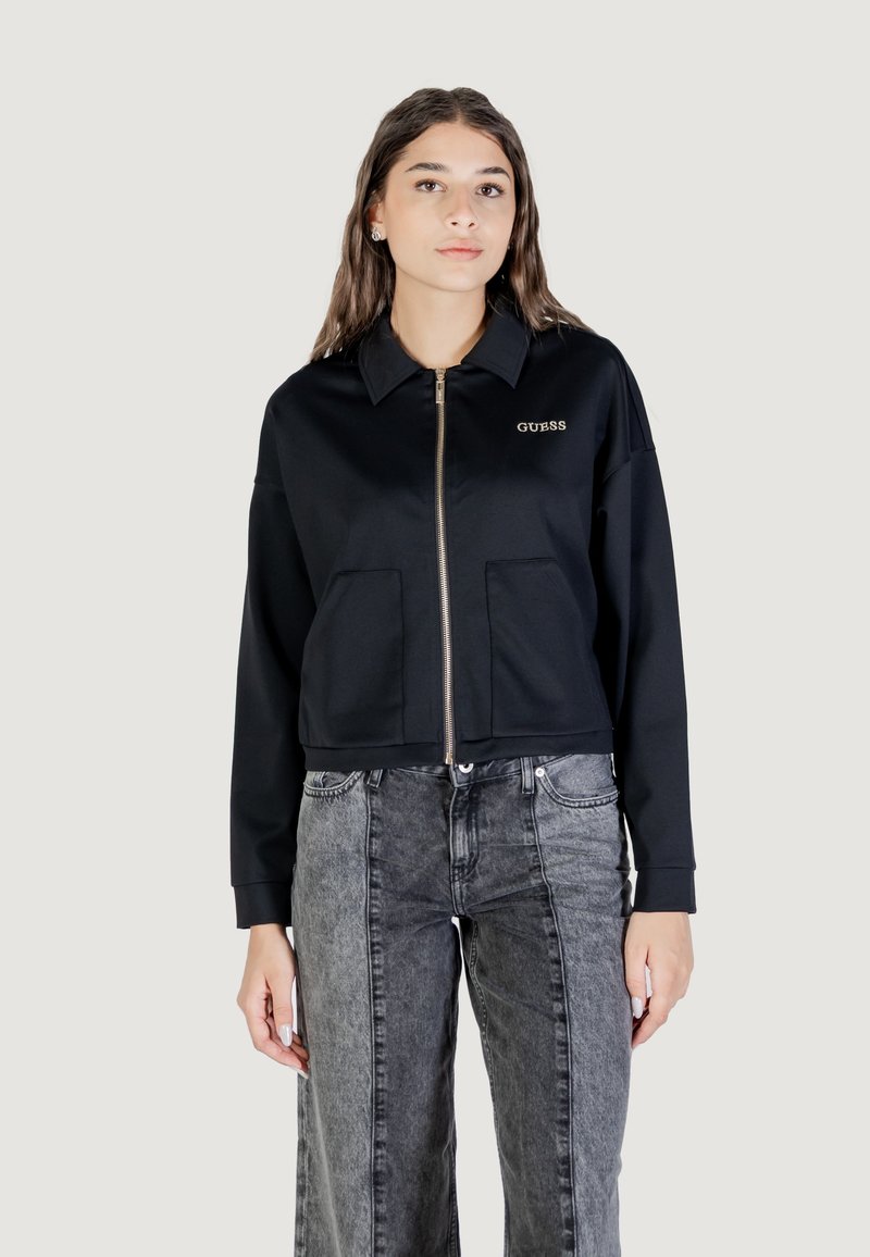 Giacca nera cropped con zip frontale, due tasche anteriori e colletto. Include il logo "GUESS" ricamato in oro sul petto.