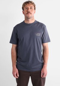 T-shirt en coton marine à manches courtes, avec un patch logo rectangulaire violet et blanc sur le côté gauche de la poitrine.