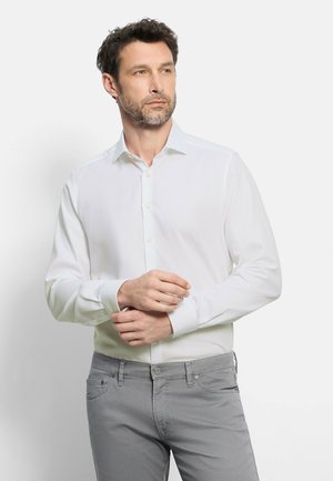 Uomo con capelli corti e scuri e barba, che indossa una camicia bianca con bottoni e pantaloni grigi, che sistema il polsino sinistro su uno sfondo neutro.