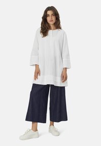 Witte long-sleeve tuniek met knoopsluiting en geborduurde accenten bij de manchetten, gecombineerd met marineblauwe wijde broek en witte sneakers.