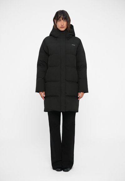 Doudoune noire avec capuche, au design carré, texture matelassée épaisse, et un petit logo sur la poitrine. Associée à un pantalon noir.