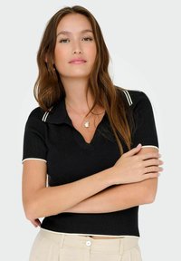 Čierny rebrovaný polo top s krátkym rukávom a výstrihom do V s bielym pruhovaným golierom a okrajmi rukávov, nosený s béžovými nohavicami.