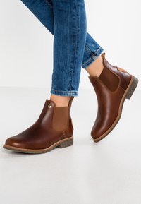 Botas de tobillo de cuero marrón con paneles laterales elásticos, tacón marrón texturizado y un acento beige. Las suelas son planas y están hechas de un material duradero.