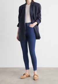 Blazer de gran tamaño en color navy, top ajustado blanco, jeans skinny azul oscuro y mules beige sobre un fondo neutro, exhibiendo un tejido elegante y un diseño moderno.