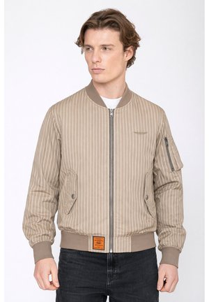 Junger Mann trägt beige Bomberjacke mit Nadelstreifen, Reißverschlusstaschen und gerippten Bündchen, kombiniert mit schwarzen Jeans, schaut nach links.