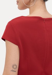 Rød kjole med korte ermer, glatt tekstur, glidelåsdetalj bak, rund hals og en tettsittende silhuett som fremhever midjen.
