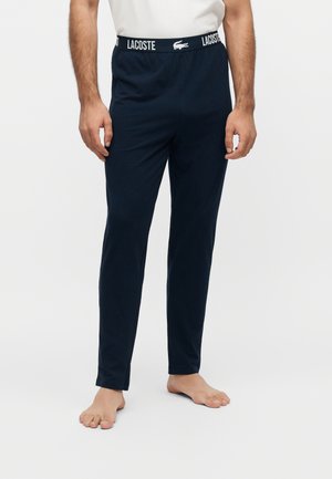 Homme portant un pantalon de détente Lacoste bleu marine avec une ceinture élastique de marque, debout pieds nus sur un fond clair uni.