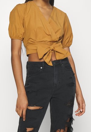 Frau trägt senfgelbes Wickel-Crop-Top mit Puffärmeln und gebundener Schleife, kombiniert mit schwarzen hoch taillierten, zerrissenen Jeans.
