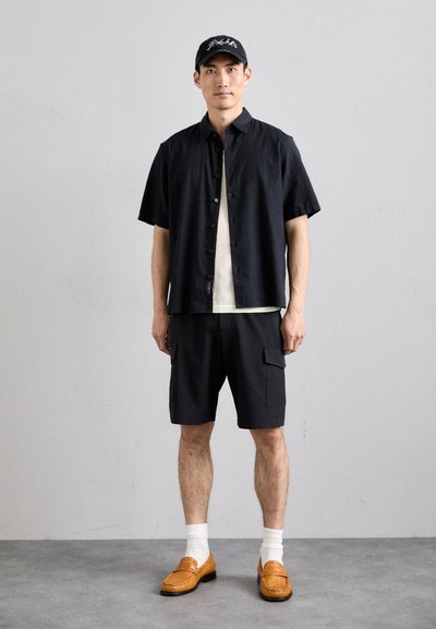 Čierna krátkovesová košeľa cez krémovú košeľu, v kombinácii s čiernymi cargo šortkami a oranžovými pletenými poltopánkami. Biele ponožky dokončujú vzhľad.