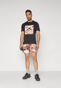 Svart T-skjorte med grafisk design, kombinert med flerfargede shorts med teksturert mønster. Grå sokker og svarte treningssko.