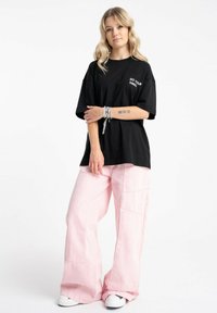 Czarny bawełniany oversize'owy T-shirt z białym napisem "DO YOUR THING" na klatce piersiowej, zestawiony z jasnoróżowymi spodniami o szerokich nogawkach i białymi trampkami.