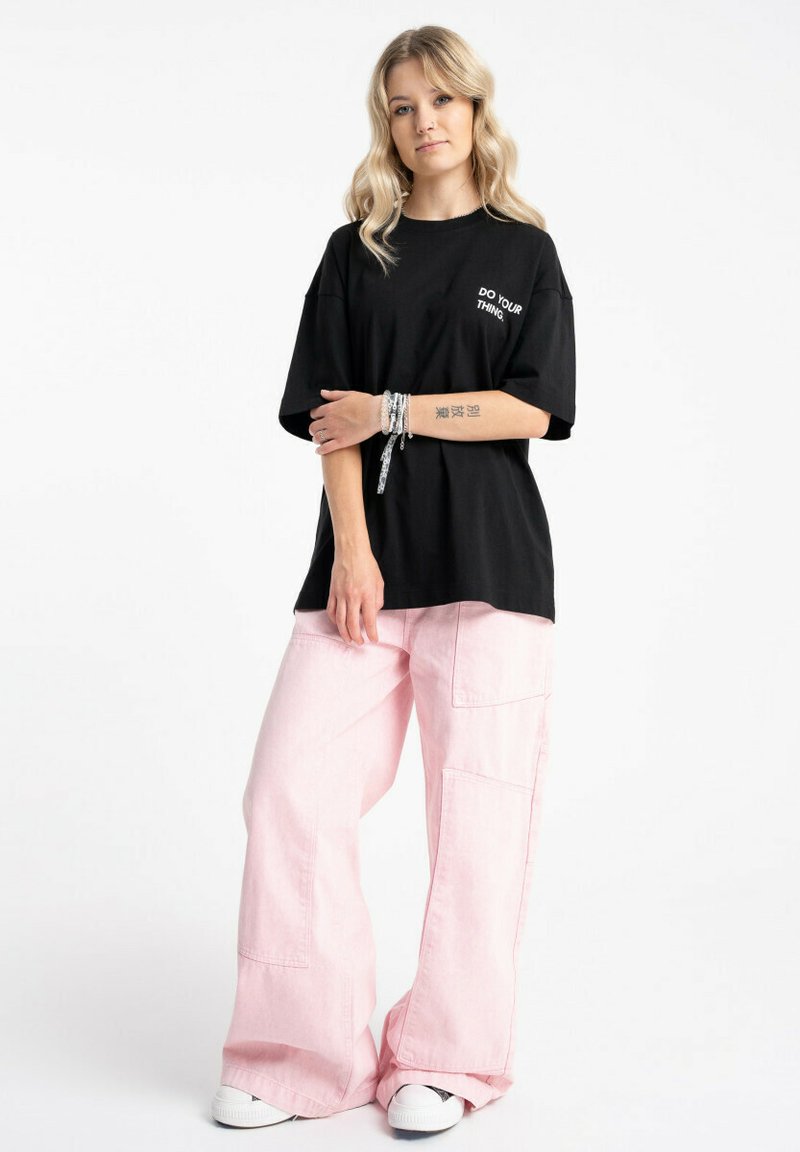 Czarny bawełniany oversize'owy T-shirt z białym napisem "DO YOUR THING" na klatce piersiowej, zestawiony z jasnoróżowymi spodniami o szerokich nogawkach i białymi trampkami.