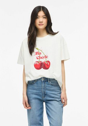 Jeune femme aux longs cheveux foncés portant un T-shirt blanc avec des cerises rouges et le texte « Ma Chérie », associé à un jean bleu.