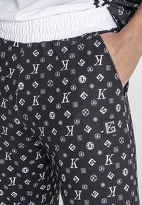 Zwarte broeken met een herhalend wit logopatroon. Voorzien van een elastische tailleband, zijzak en een klein zwart labelaccent.