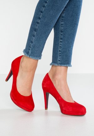 Piernas con jeans azules de dobladillo deshilachado y elegantes zapatos de tacón alto de ante rojo brillante sobre un suelo blanco.
