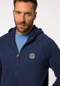 Námořnická zipová mikina s kapucí, s lehkým modrým vnitřním podšitím. Obsahuje obdélníkovou logo nášivku na hrudi a texturovanou tkaninu.
