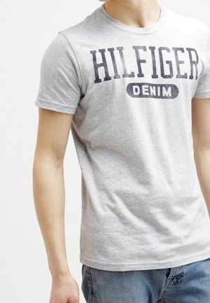 Persoon draagt een lichtgrijs T-shirt met "HILFIGER DENIM" in donkere letters gedrukt over de borst en een blauwe spijkerbroek.