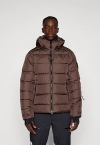 Piumino marrone con motivo trapuntato verticale, cappuccio, cerniera frontale e tasche laterali. Presenta polsini elastici e dettagli in metallo.