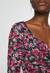 Femme portant une robe noire à motif floral rose et vert et une boucle d'oreille argentée pendante, montrée de l'épaule à la bouche.