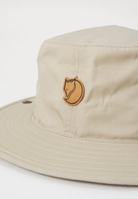 Chapeau seau beige en tissu léger, avec un logo de renard brun brodé sur le côté et une large visière plate.