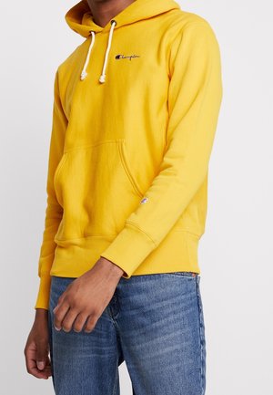 Man met een felgele Champion-hoodie met witte trekkoorden en een blauwe spijkerbroek, staand tegen een effen witte achtergrond.