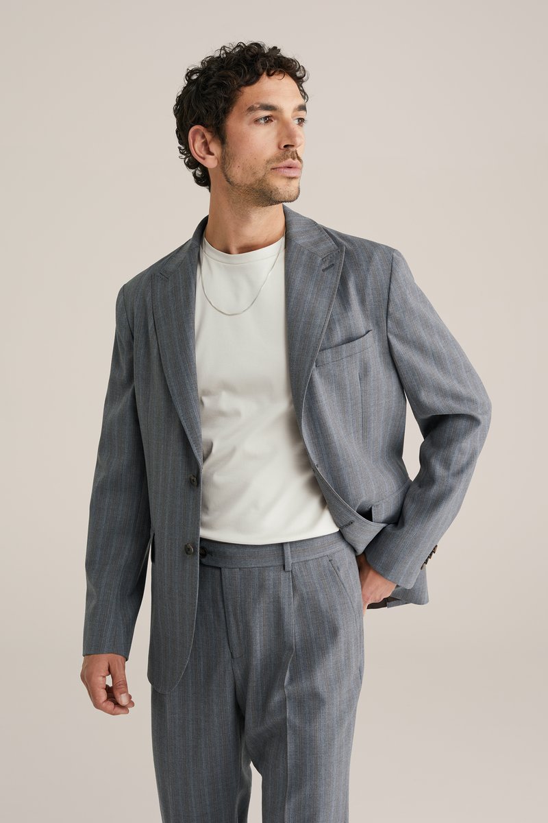 Homme aux cheveux bouclés portant un costume bleu à fines rayures et une chemise blanche, posant avec une main dans la poche, regardant sur le côté.