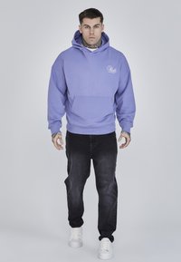 Sudadera con capucha color lavanda hecha de tela suave, con un bolsillo frontal. Combinada con jeans oscuros y zapatillas blancas. Detalle de logo sencillo en el pecho.