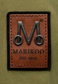 Ploská, obdĺžniková kožená nášivka v hnedej farbe s embosovaným strieborným logom "M" a textom "MARIKOO EST 2016". Čierny okraj z filcu; khaki pozadie.