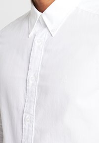 Camisa de algodón blanca con botones que presenta un cuello clásico, una tapeta de doble capa y un sutil logo bordado en el área del pecho izquierdo.