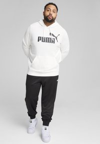 Hanorac alb cu glugă, având logo-ul „PUMA” în negru, asortat cu pantaloni jogger negri și adidași albi. Material moale, croi casual, design simplu.