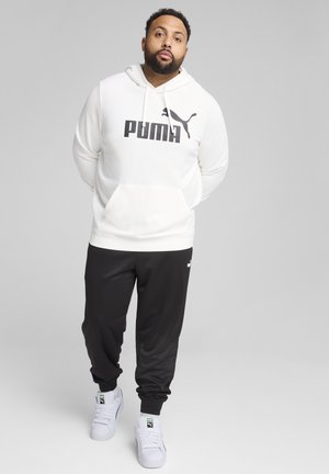 Biała bluza z kapturem z czarnym logo „PUMA”, zestawiona z czarnymi dresami i białymi butami sportowymi. Miękki materiał, swobodny krój, prosty design.
