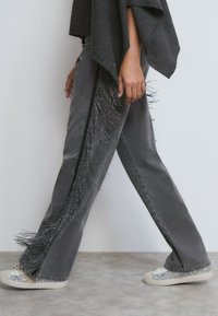 Persona caminando con vaqueros grises con flecos negros a los lados, combinados con zapatillas plateadas brillantes y una camiseta gris oscuro.