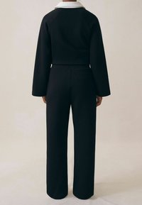 Čierny overal s dlhým rukávom a bielym golierom, vo voľnom strihu, so bočnými vreckami, vyrobený z hladkého, textúrovaného materiálu.