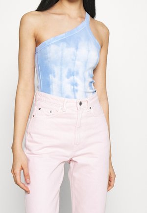 Kvinde iført en blå og hvid enskulder tie-dye top med højtaljede lys rosa bukser, stående mod en hvid baggrund.