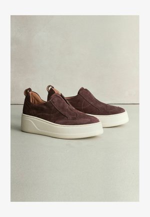 Hnědé semišové slip-on boty mají polstrovanou bílou podrážku, hladký povrch a viditelné švy. Zadní poutko pro snadné obouvání.