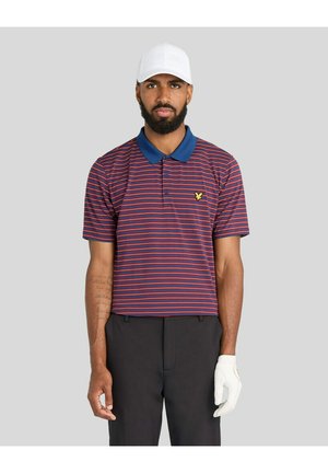 Mand iført en rød og blåstribet poloshirt, sorte bukser, hvid golfhandske på venstre hånd og en hvid kasket, stående mod en grå baggrund.