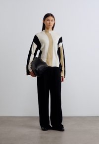 Pull tricoté en crème, noir et beige, avec un motif géométrique ; associé à un pantalon large noir et un sac noir.