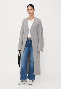 Lang grå cardigan laget av mykt materiale, åpen front, oversized ermer, sammen med en hvit crop top og blå flagrende jeans. Svart tilbehør.