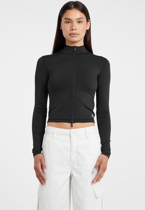 Veste noire courte zippée avec manches longues, fabriquée en tissu extensible lisse. Elle présente un col montant et un design ajusté.