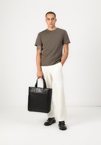 Schwarze Leder-Tote-Bag mit doppelten Griffen, glatter Textur, rechteckiger Form und minimalistischem Design, getragen von einer Person in einem neutralen Outfit.