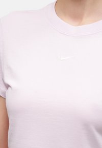 Γυναίκα φοράει ένα ανοιχτό ροζ μπλουζάκι Nike με στρογγυλή λαιμόκοψη και μικρό λευκό λογότυπο Nike στο στήθος.