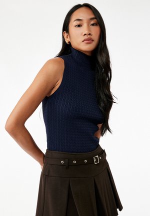 Marineblauwe geribbelde trui zonder mouwen met hoge hals, gecombineerd met een bruine geplooide rok met een gespsluiting en decoratieve hardwareaccenten.