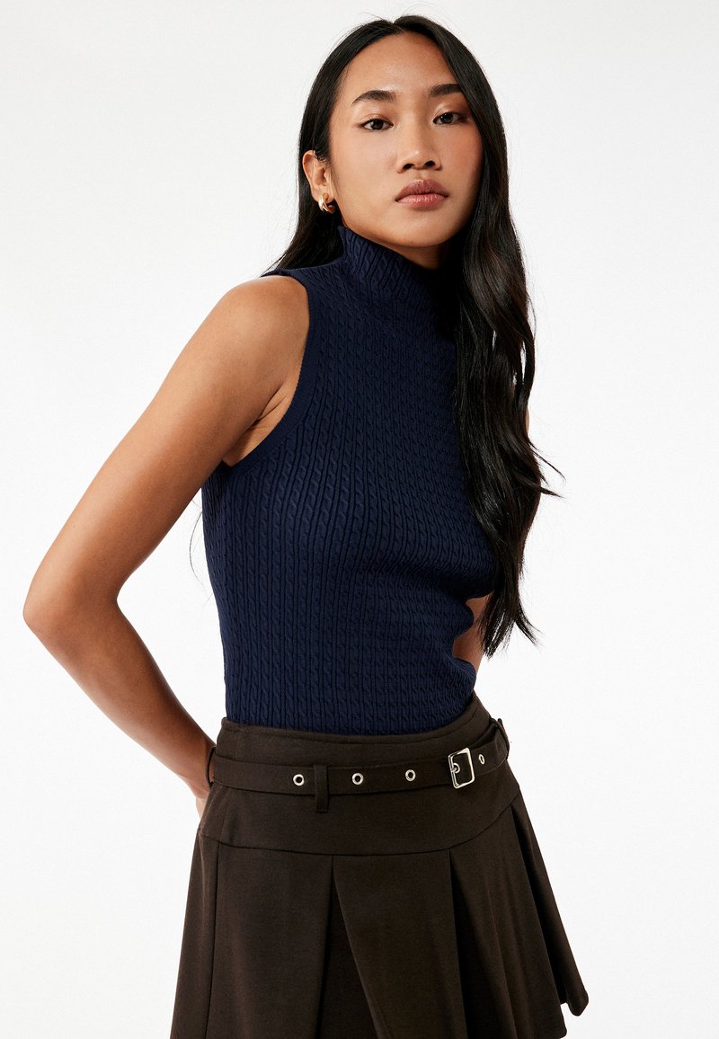 Suéter sin mangas de cuello alto de canalé en color navy, combinado con una falda plisada marrón que cuenta con un cinturón con hebilla y detalles decorativos de hardware.