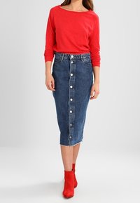 Punainen pitkähihainen paita, joka on yhdistetty polvimittaiseen farkkuhameeseen, jossa on etunappikiinnitys ja lievää kulumista helman kohdalla. Punaiset nilkkurit.