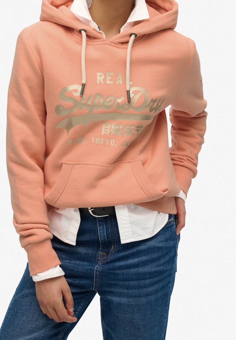 Personne portant un sweat à capuche couleur pêche avec le logo "SuperDry", chemise blanche en dessous, jean bleu et ceinture noire, les mains reposant près des poches.