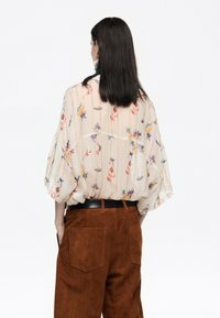 Blusa transparente de estampado floral en color beige claro con rayas verticales sutiles, combinada con pantalones de ante marrón de tiro alto con bolsillos traseros.