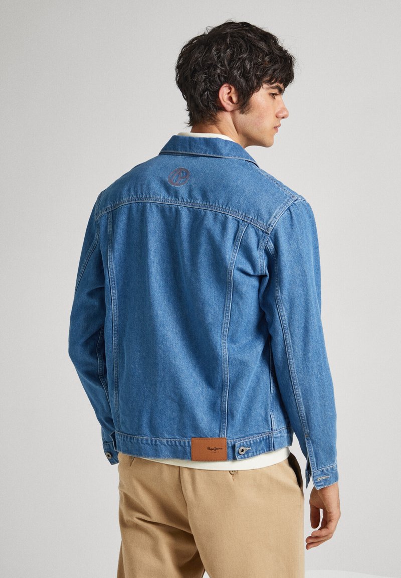 Chaqueta de mezclilla azul con un cuello estructurado, detalle de logo en la espalda y parche de cuero en el dobladillo. Mangas largas y un corte relajado.